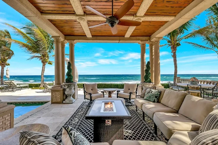 A high-end covered outdoor patio with a wood-plank ceiling and ceiling fan, featuring a modern fire pit table, plush beige seating, and a clear view of the turquoise Atlantic Ocean and white sand beach in Florida.